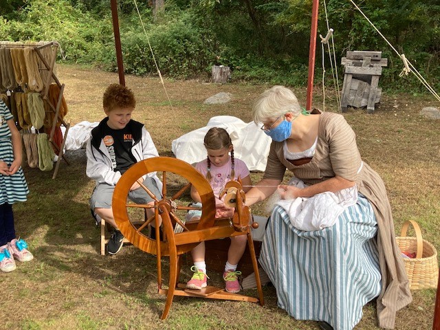 A young girl learns to spin on an 18th-century replica wheel under the guidance of a costumed instructor. 