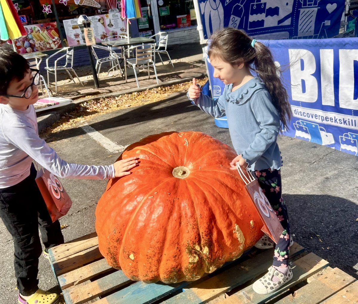 Children gather around the giant pumpkin, some trying to lift it while others pose for photos beside the bright orange vegetable.